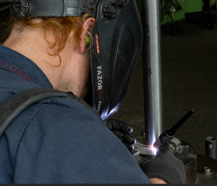 Soudeur portant un casque, soudant deux tuyaux métalliques ensemble dans un atelier.