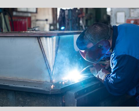 Soudeur en salopette bleue et masque soudant une structure métallique dans un atelier.
