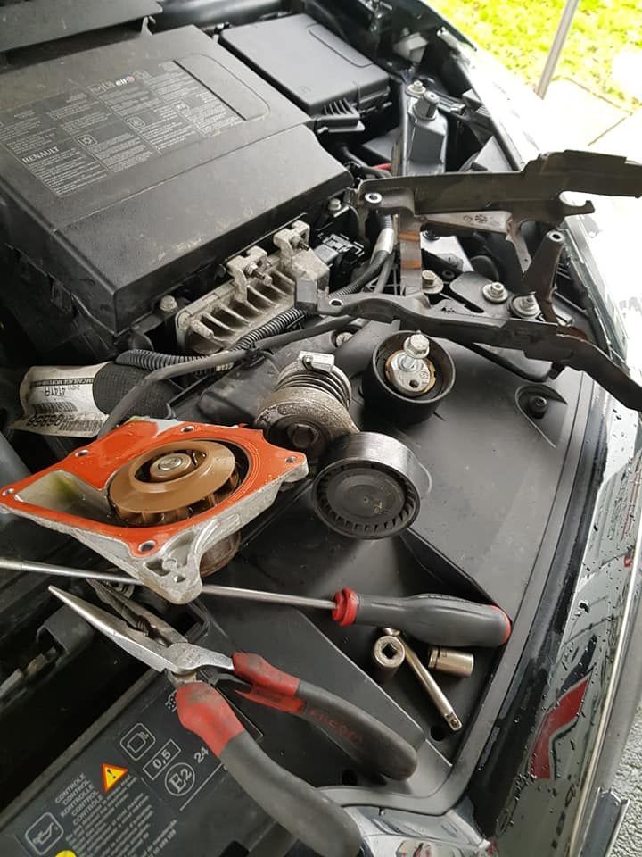 Compartiment moteur ouvert, outils et pièces démontées visibles : pompe à eau, poulie, tendeur de courroie et câblage.