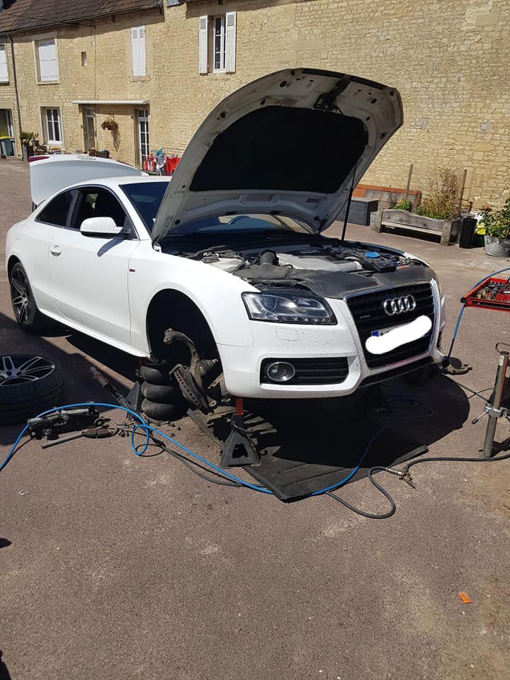 Coupé Audi blanc, capot ouvert, soulevé par un cric, roues avant démontées, en cours de réparation à l'extérieur.