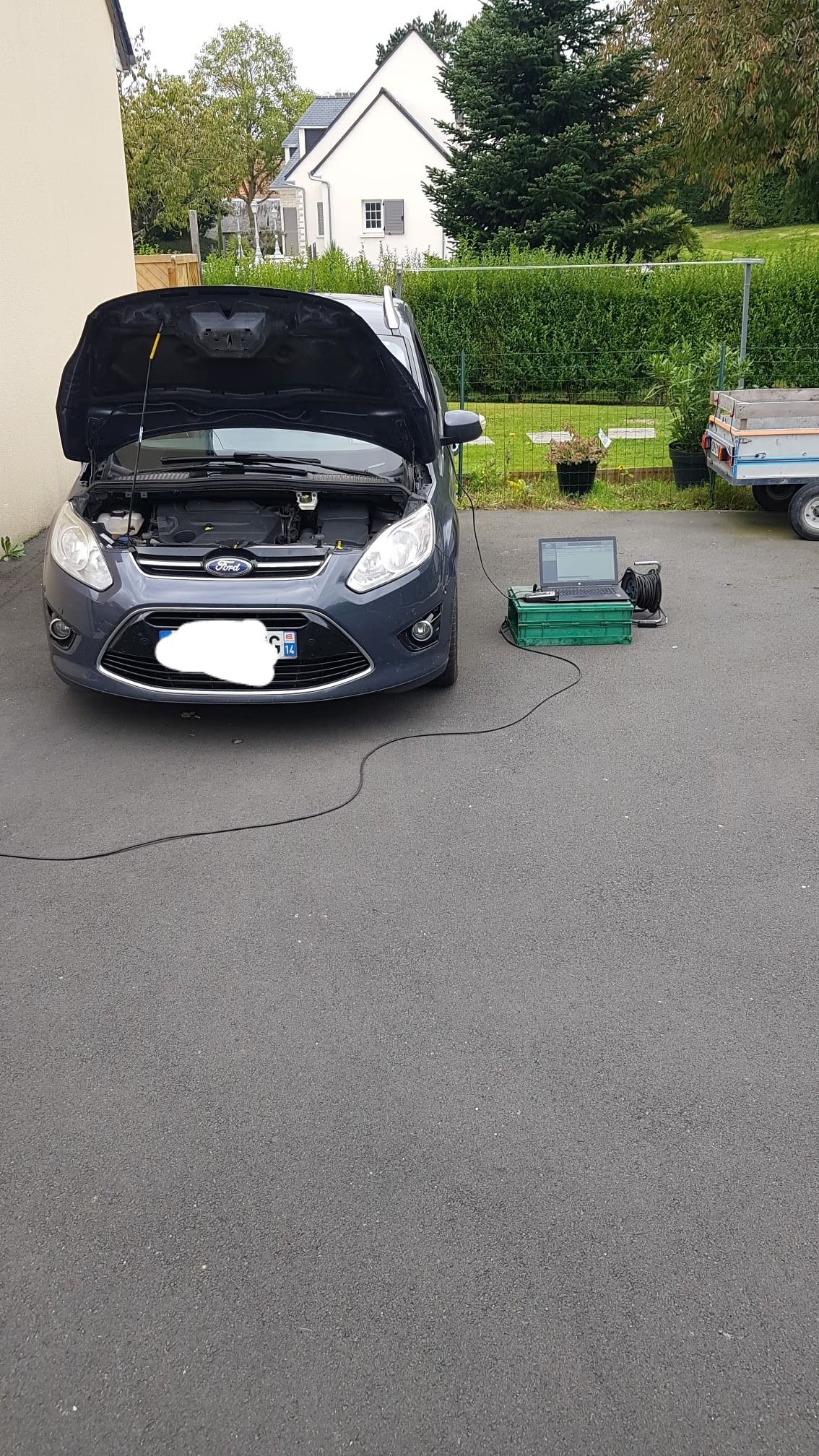 Une Ford grise, capot ouvert, branchée à un appareil de diagnostic dans une allée.