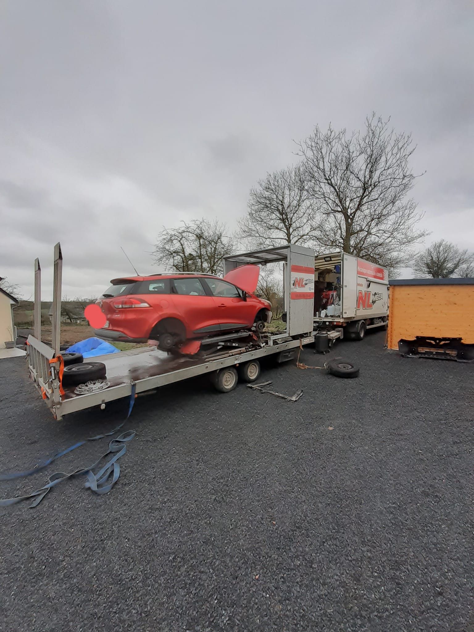 Voiture rouge sur une remorque transportée par un semi-remorque sur une surface de gravier sous un ciel nuageux.