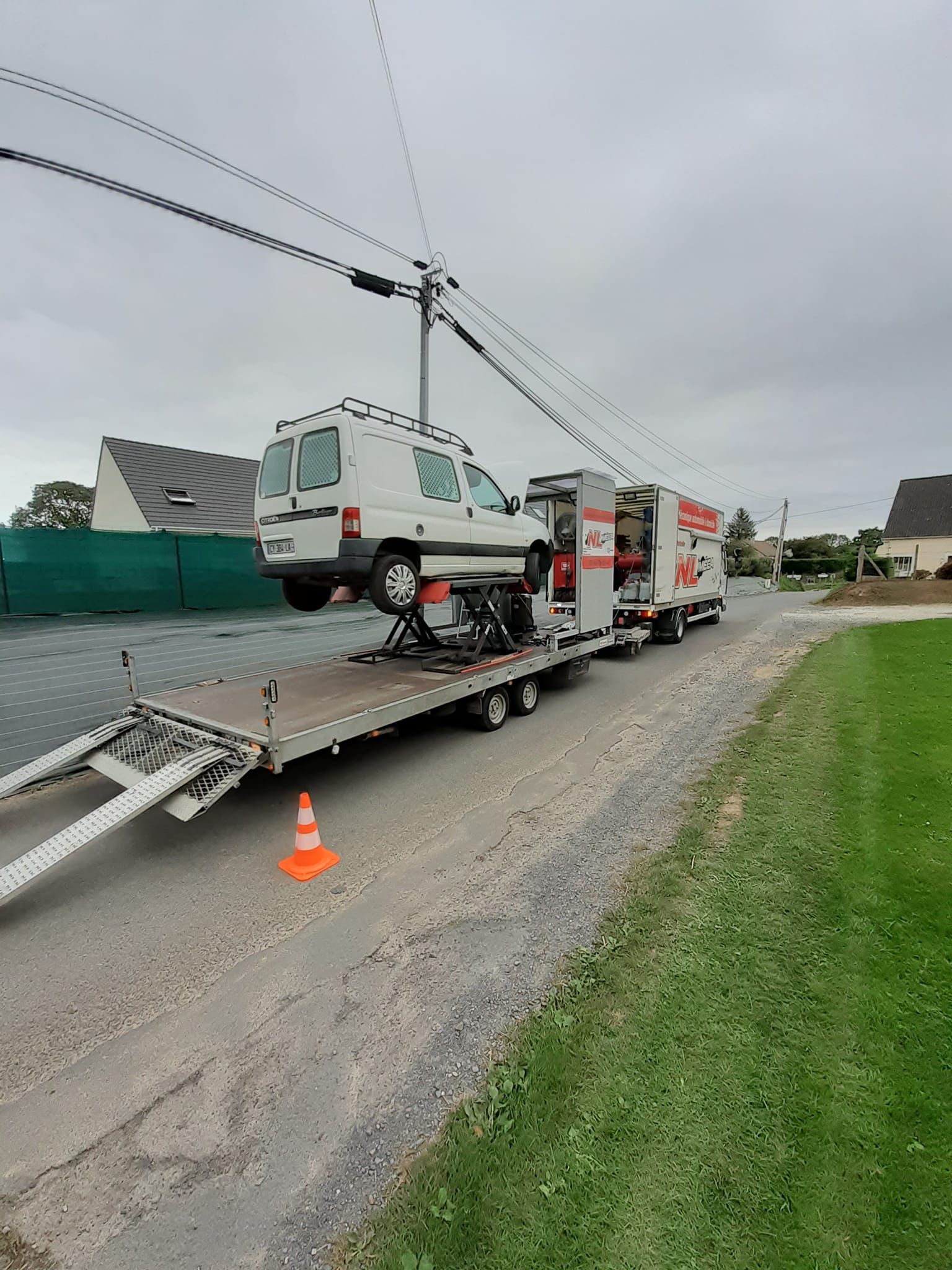 Une camionnette blanche transportée sur une remorque de dépanneuse sur une route goudronnée, sous des lignes électriques, sous un ciel nuageux.