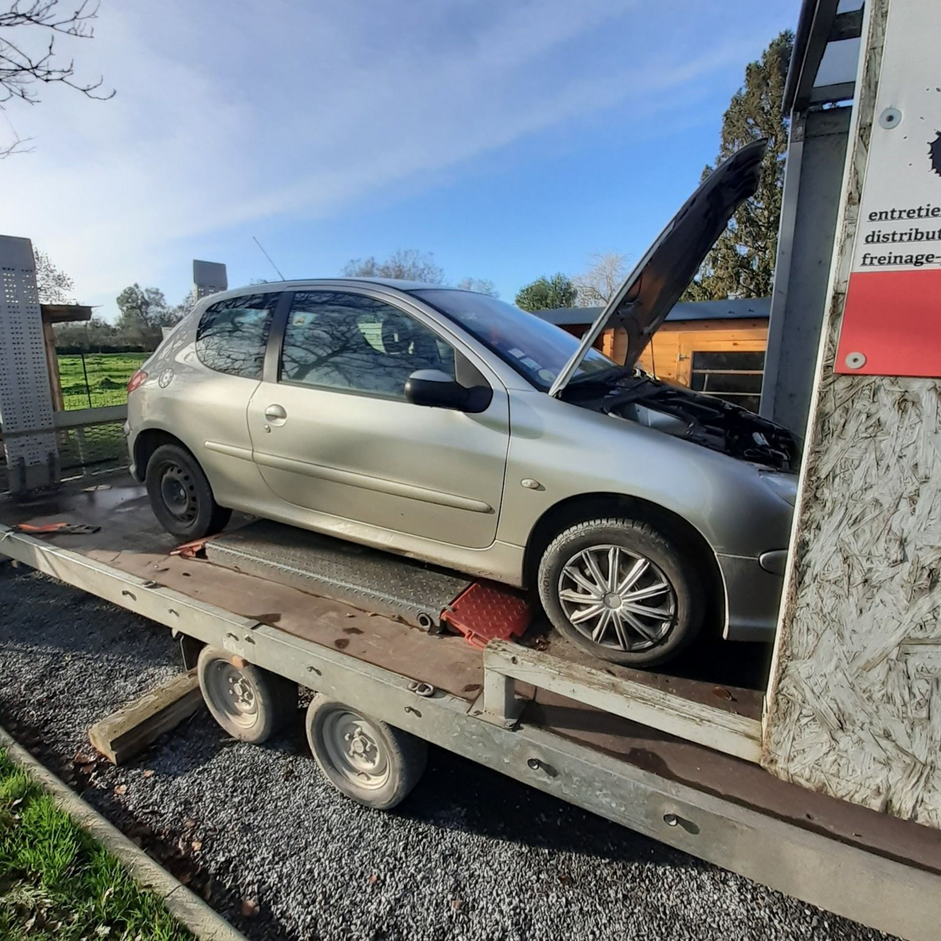 Voiture argentée, capot ouvert, sur une remorque à plateau, en extérieur.