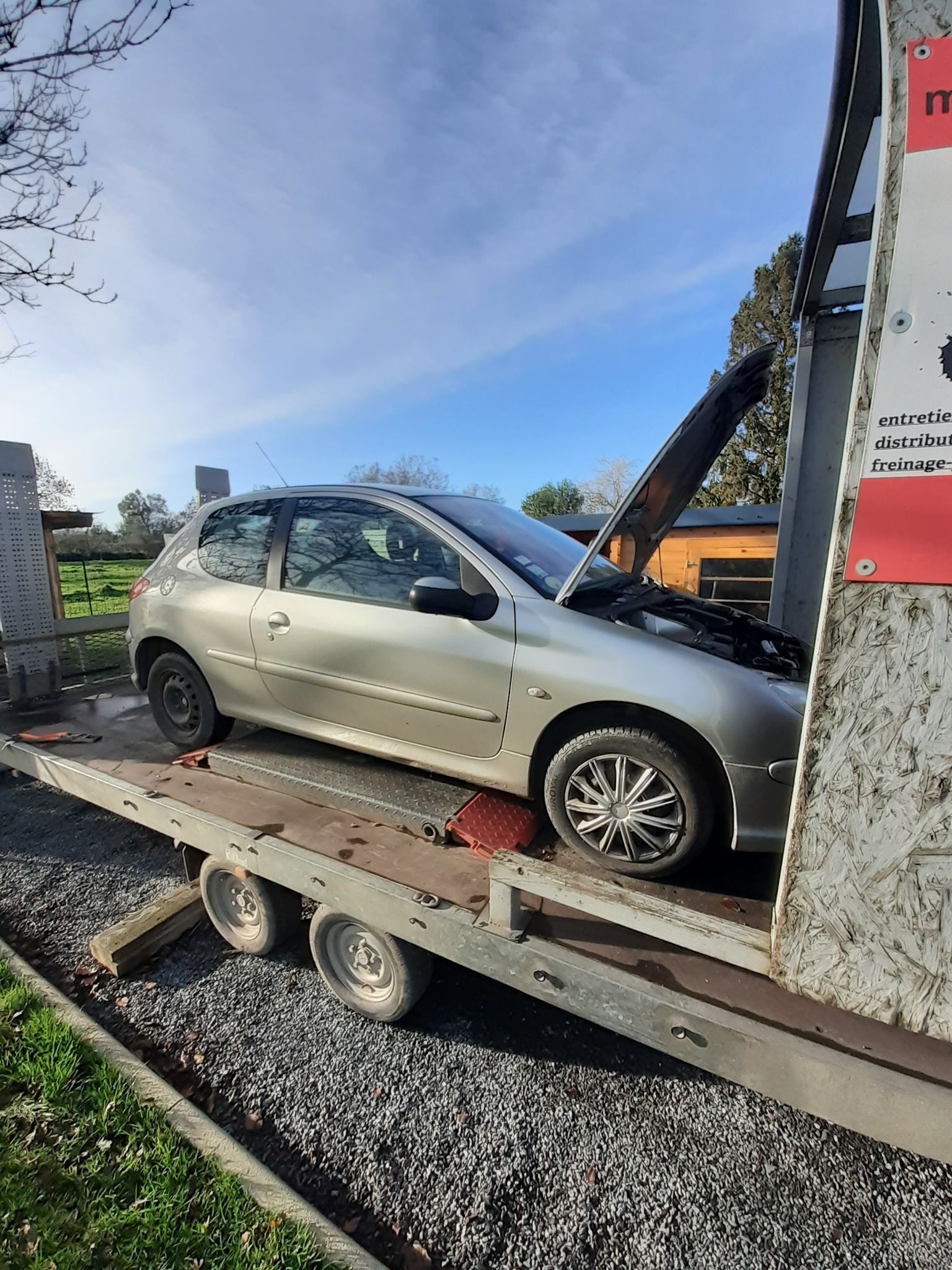 Voiture argentée sur une remorque à plateau, sous un ciel bleu. La voiture se trouve près d'une zone de gravier, en plein jour.