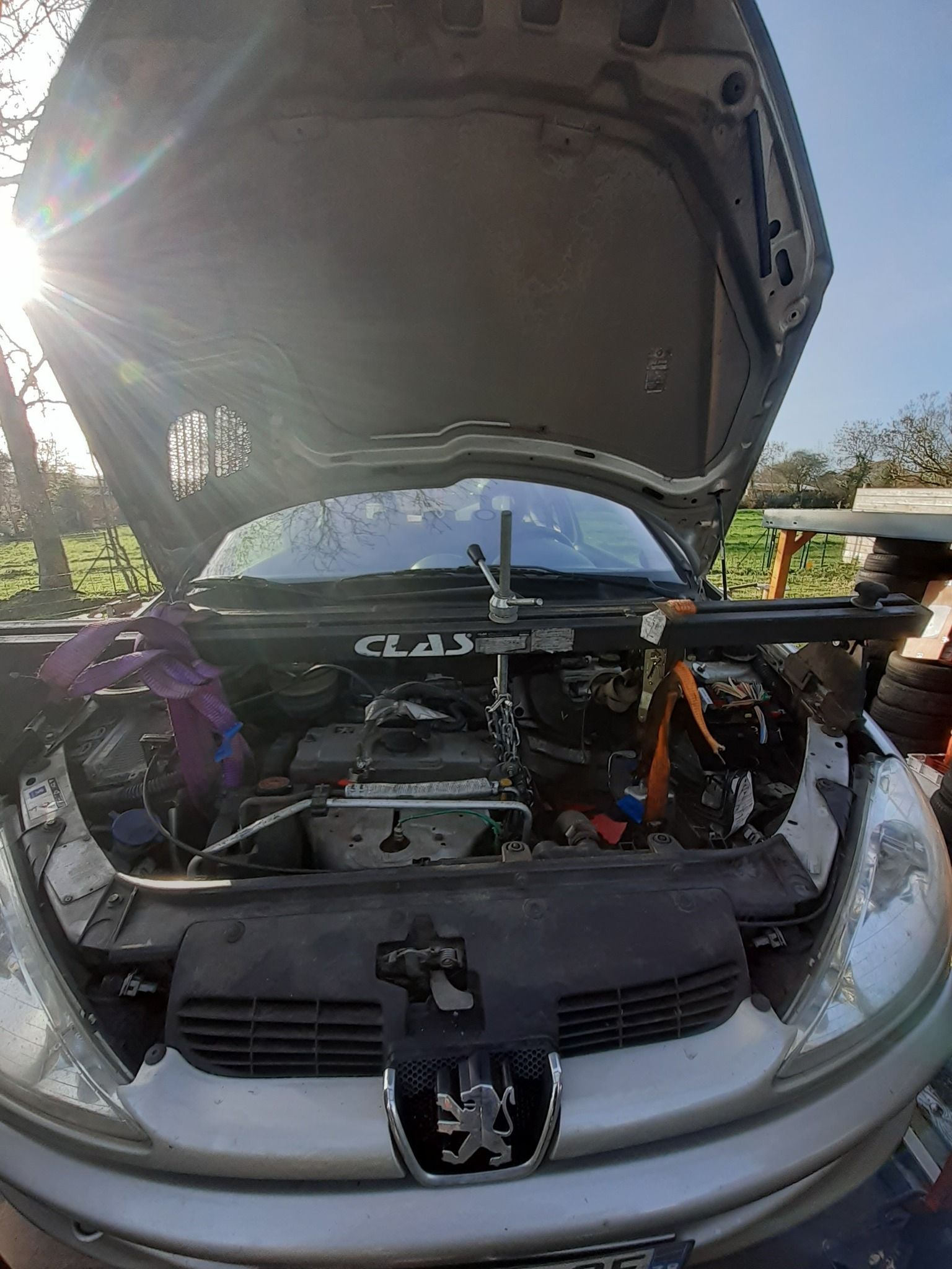 Un moteur de voiture en cours de réparation, capot ouvert, en extérieur, sous un soleil éclatant.