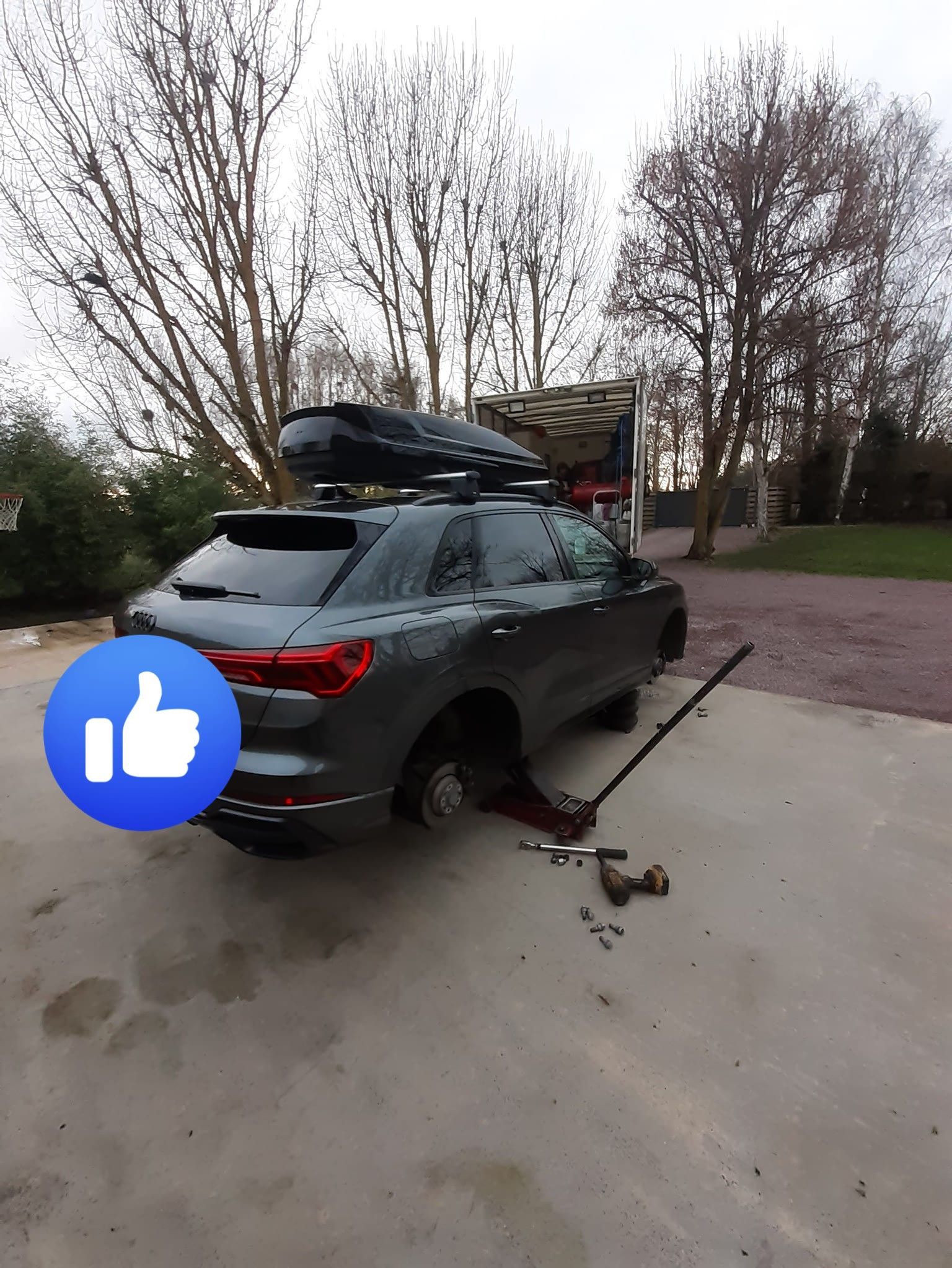 Un SUV gris avec un pneu crevé est soulevé à l'extérieur. Un emoji pouce levé est superposé à la photo.