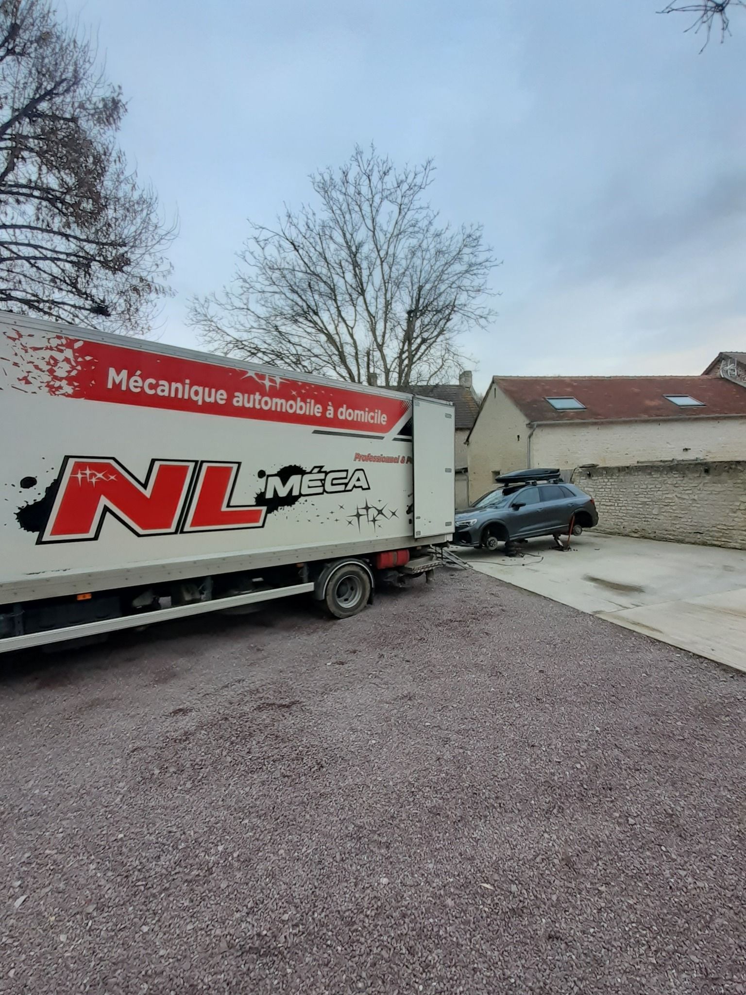 Un camion portant l'inscription NL Méca sur le côté est stationné sur du gravier à côté d'un bâtiment et d'un SUV foncé.