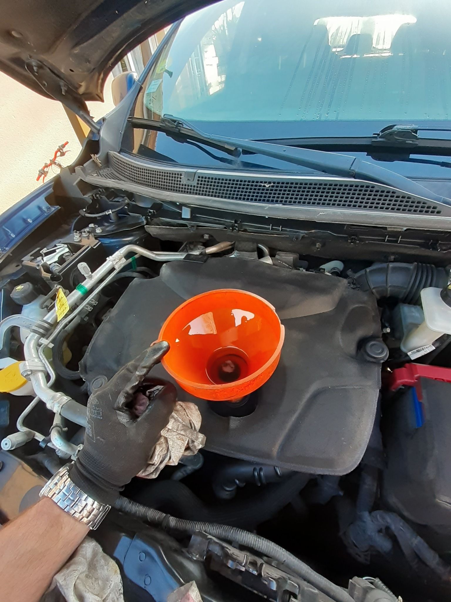 Une personne portant des gants verse un liquide à l'aide d'un entonnoir dans le moteur d'une voiture.