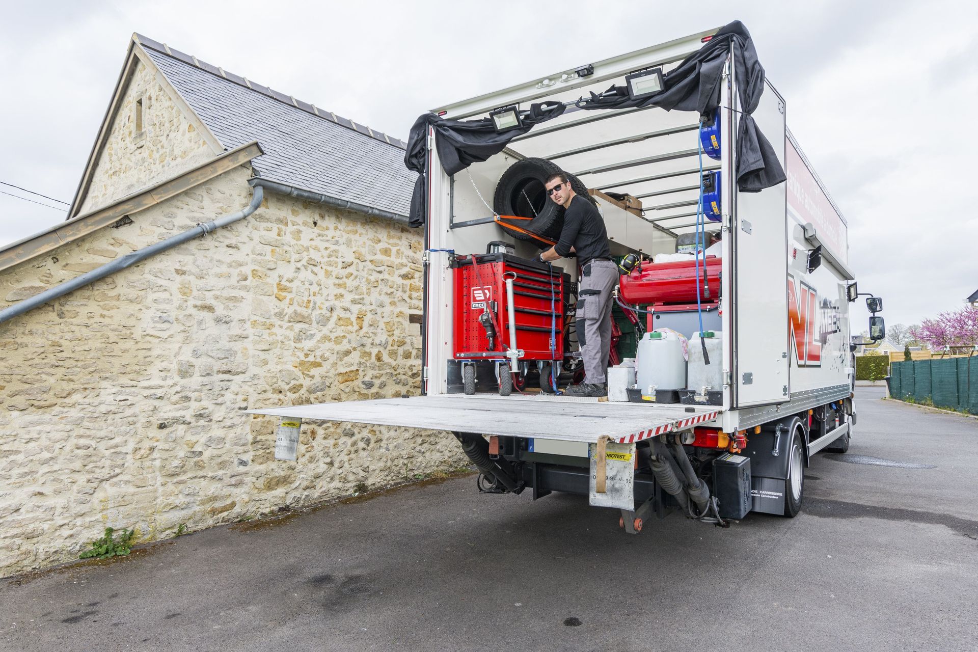 Un mécanicien travaille à l'intérieur d'un camion à plateau. On aperçoit des coffres à outils rouges et du matériel. Vue extérieure.