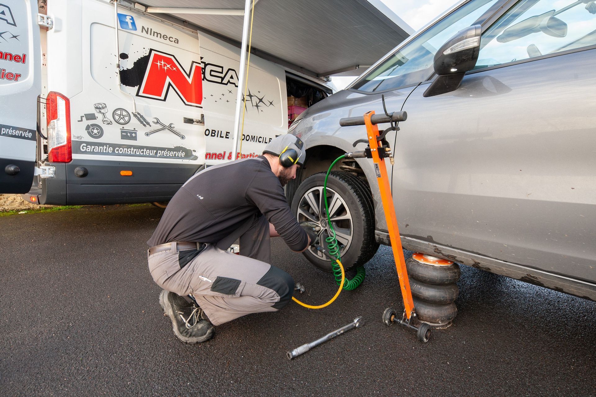 Un mécanicien gonfle un pneu de voiture près d'une camionnette ; tuyaux d'air verts et jaunes, pompe grise et orange.
