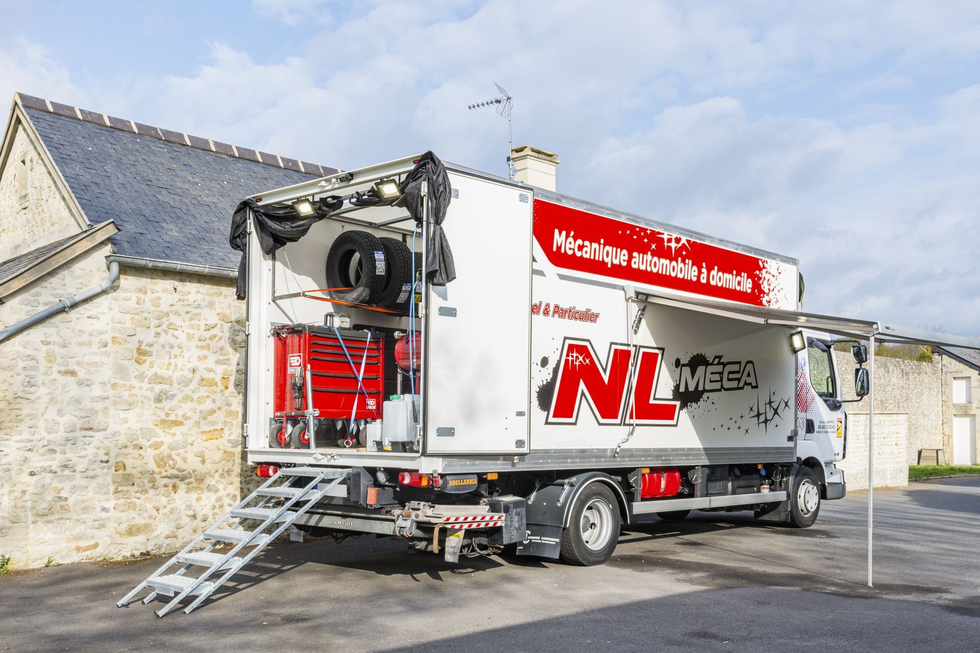Camion de mécanicien mobile avec rampes, outils et pneus stationné près d'un bâtiment en pierre.