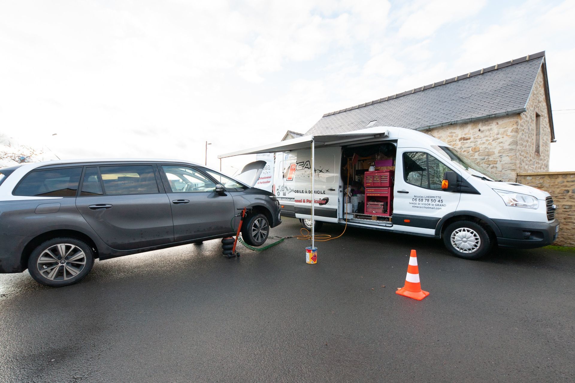 Une camionnette blanche à ridelles ouvertes est garée à côté d'une voiture grise dans une rue. Un cône orange se trouve au premier plan.