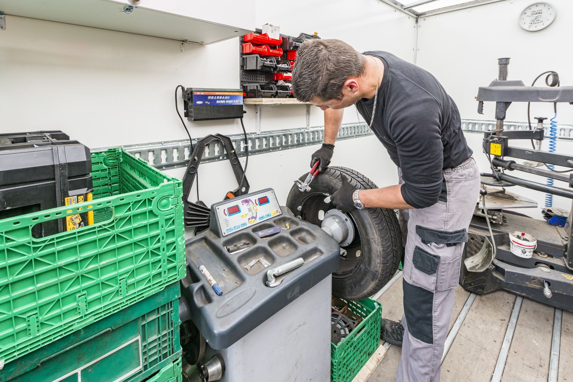 Un mécanicien équilibre un pneu dans un atelier mobile. On aperçoit des caisses vertes, des outils et une machine d'équilibrage.