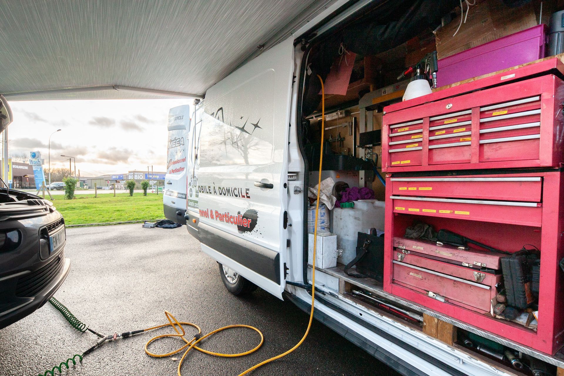 Camionnette d'assistance technique avec porte ouverte, caisse à outils rouge et voiture en réparation. À l'extérieur, sous un auvent.