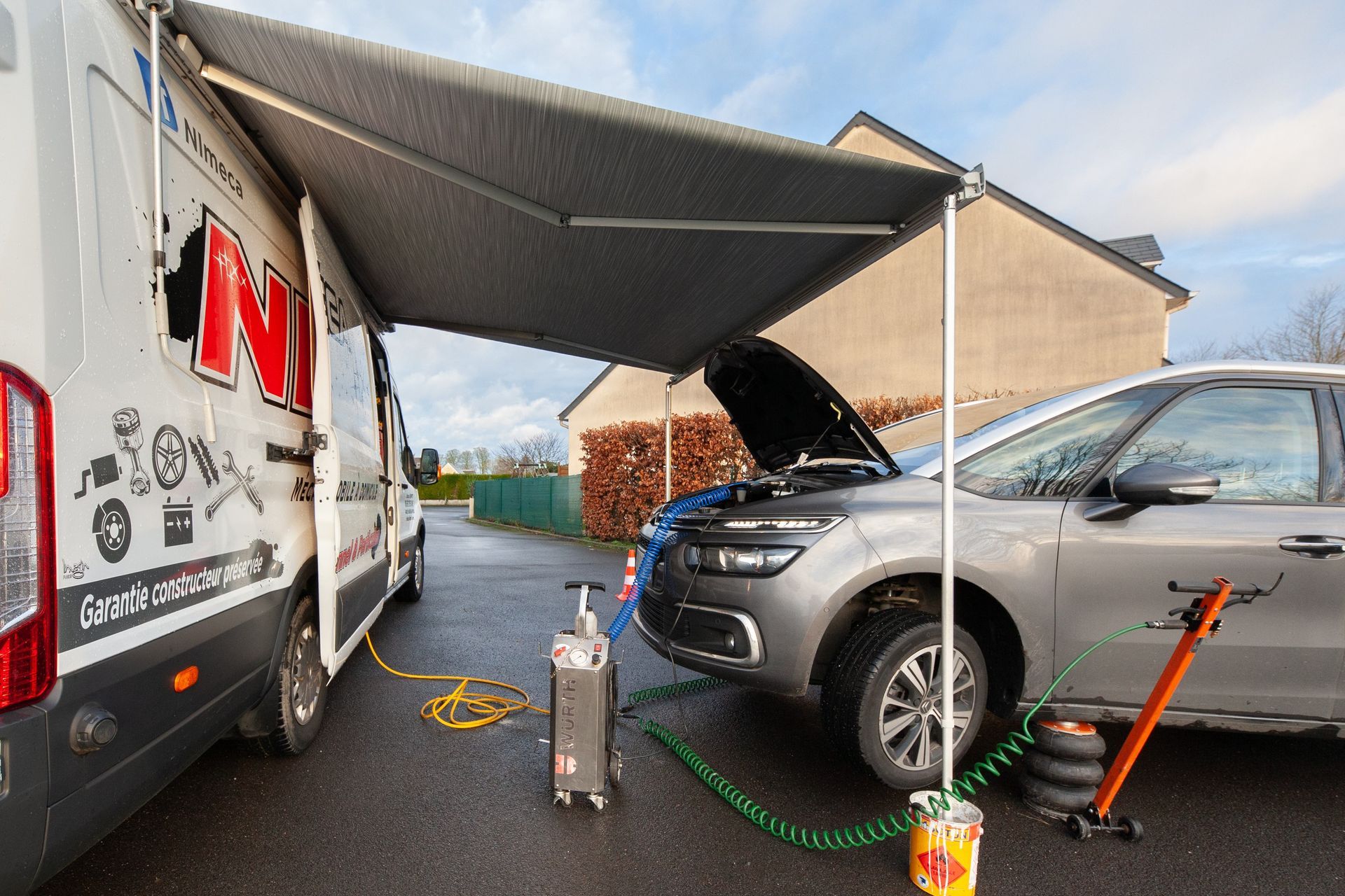 Réparation automobile mobile : fourgonnette avec auvent, capot ouvert, outils et équipements, journée nuageuse.