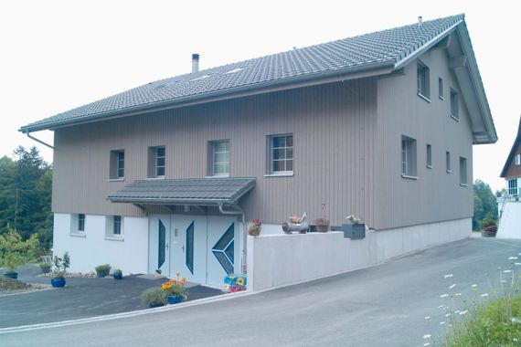 Sonnendurchflutetes Schlafzimmer mit ungemachtem Bett, grossen Fenstern und einer Holzkommode auf der rechten Seite.