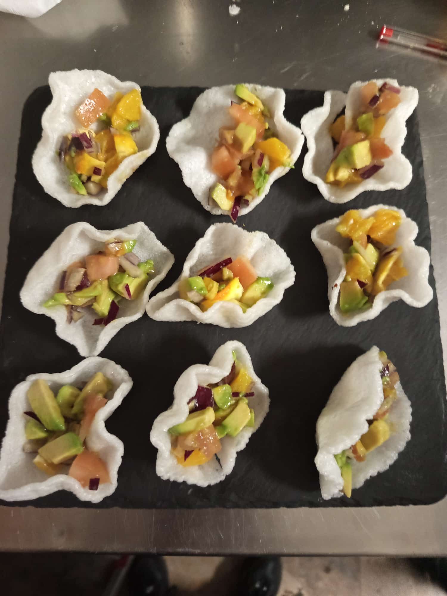 Nueve pequeños aperitivos de camarones y aguacate en vasitos de concha blanca, dispuestos sobre una bandeja oscura.