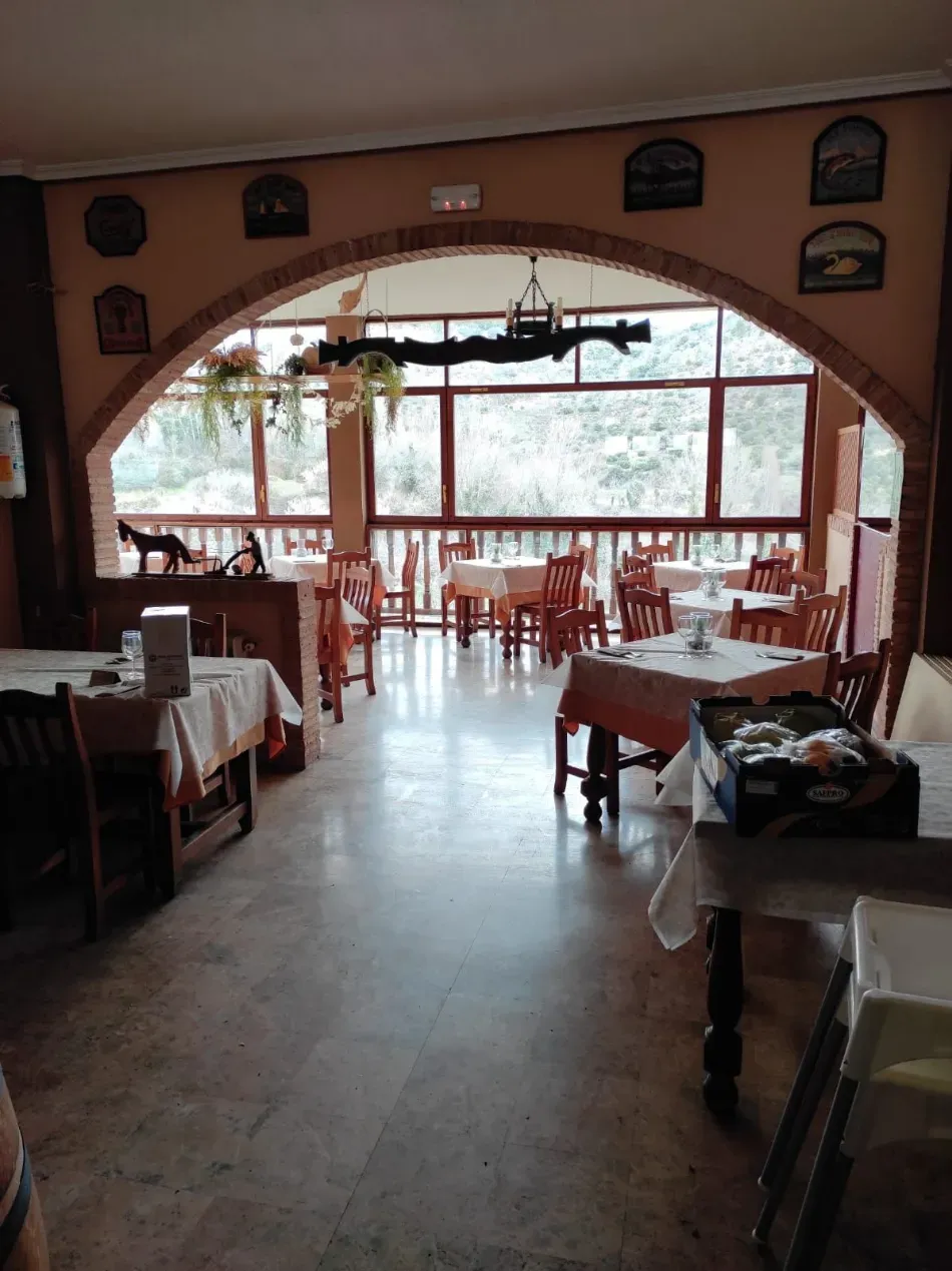 Comedor vacío de un restaurante con manteles rojos y una gran ventana arqueada con vistas a los árboles.