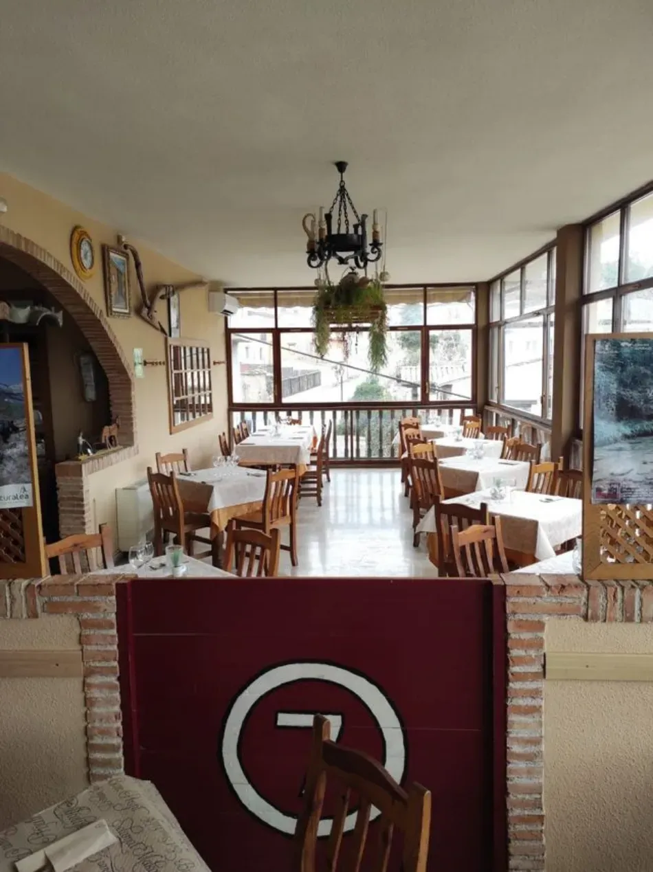 Acogedor comedor con mesas y sillas de madera, chimenea de ladrillo y grandes ventanales con vistas al exterior.
