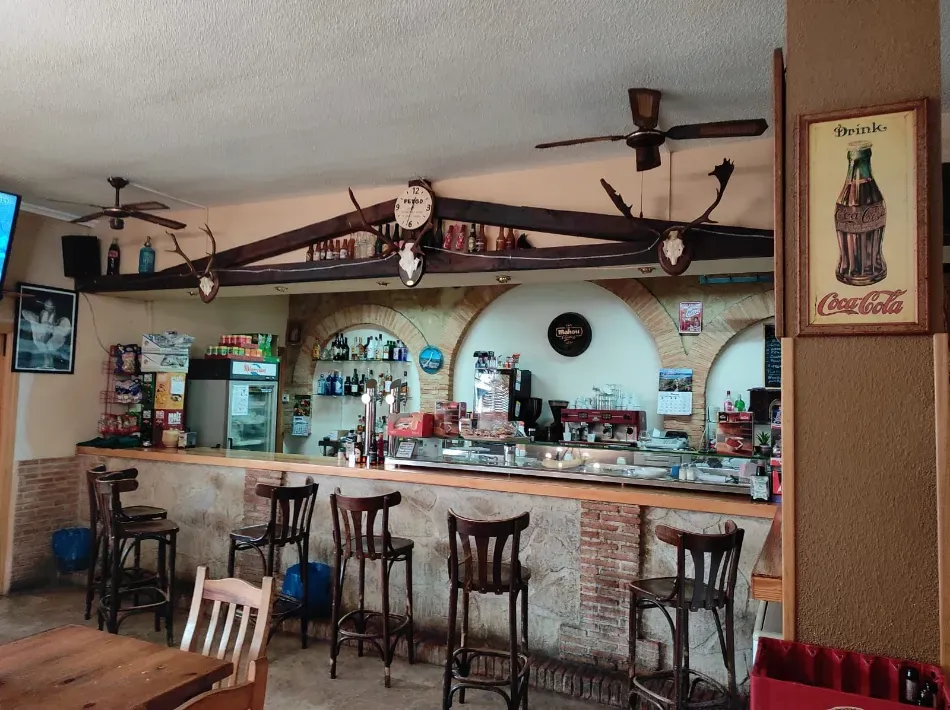 Pequeño bar de estilo rústico con taburetes, estantes con botellas y una barra con espejo bajo ventiladores de techo.