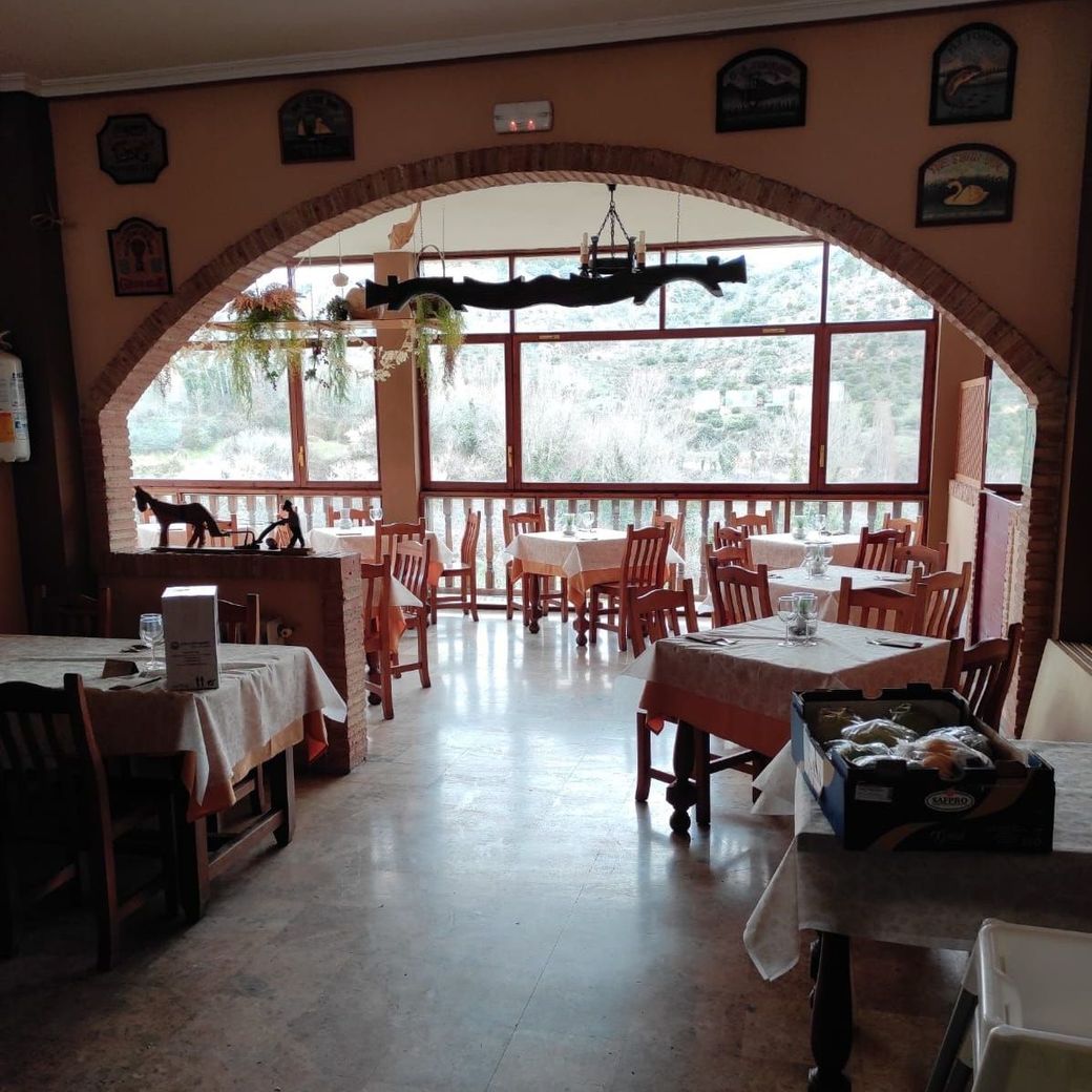 Comedor vacío de un restaurante con mesas y sillas de madera, una gran ventana arqueada y vista exterior nevada.