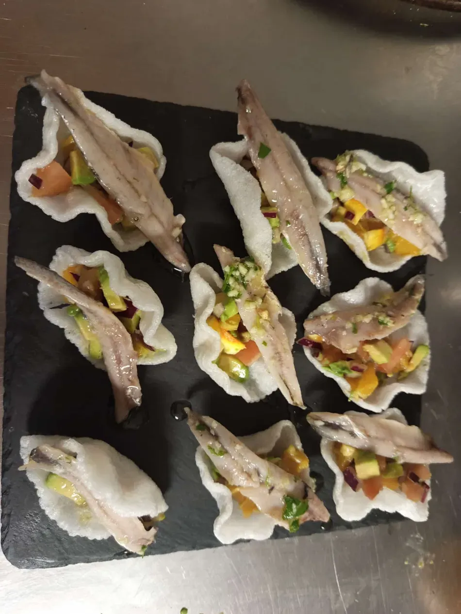 Plato de canapés variados de verduras y carne sobre galletas blancas con forma de cuchara en una bandeja de pizarra oscura.