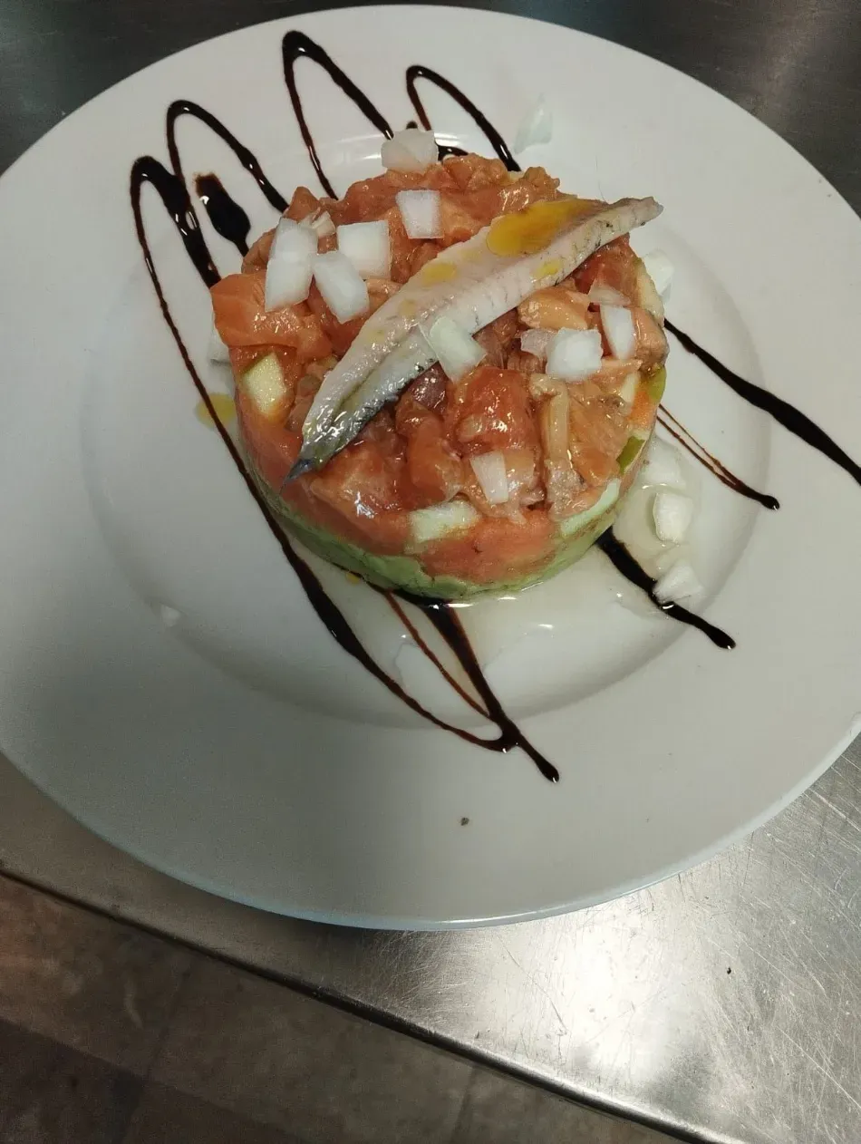 Tartar de atún gourmet en plato blanco con un chorrito de vinagre balsámico oscuro, cubierto con verduras picadas y una guarnición de pescado.