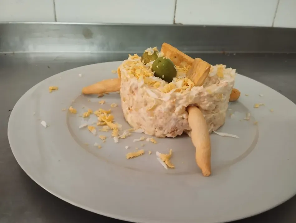 Ensalada de arroz en un tazón blanco sobre un plato, con queso rallado y una guarnición de pepinillos, sobre una encimera.