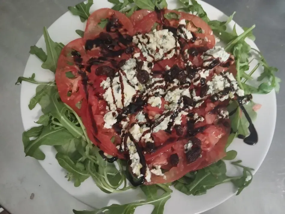 Ensalada de rúcula con rodajas de tomate, queso desmenuzado y un chorrito de vinagre balsámico en un plato blanco.