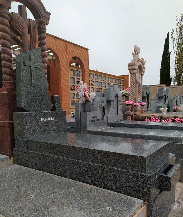 Cementerio con lápidas de granito y una cruz, con edificios de ladrillo al fondo. Flores rosas adornan algunas tumbas.