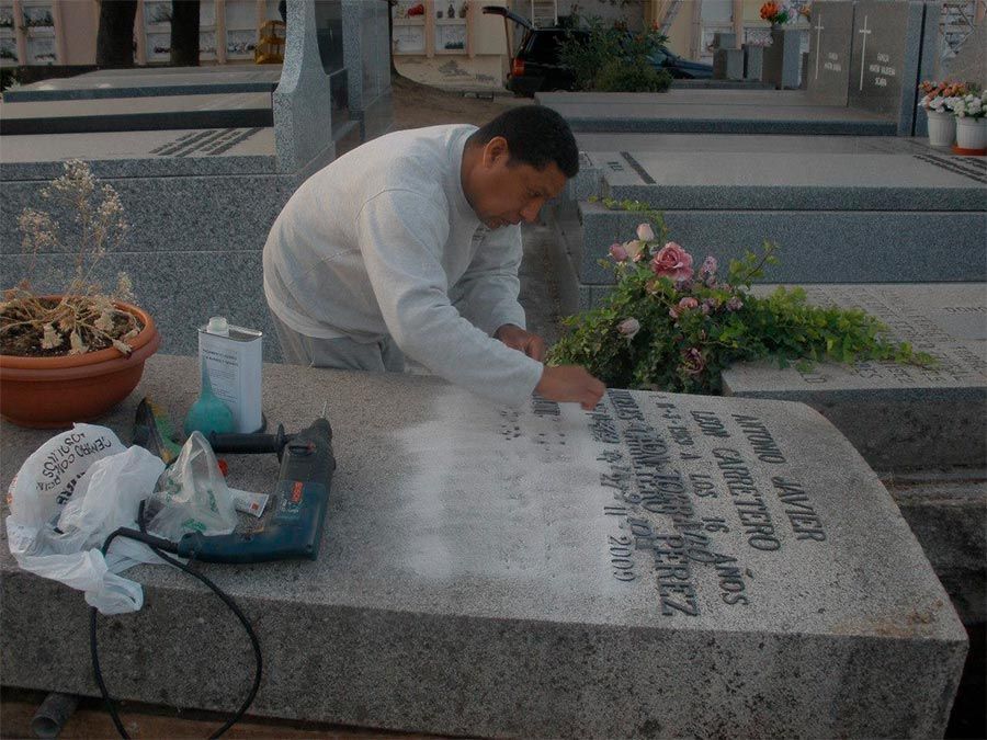 Hombre limpiando una lápida en un cementerio; flores y herramientas cerca.