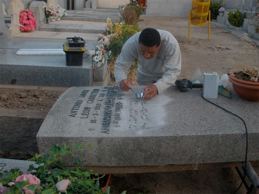 Hombre limpiando una lápida en un cementerio con herramientas y flores a la vista.