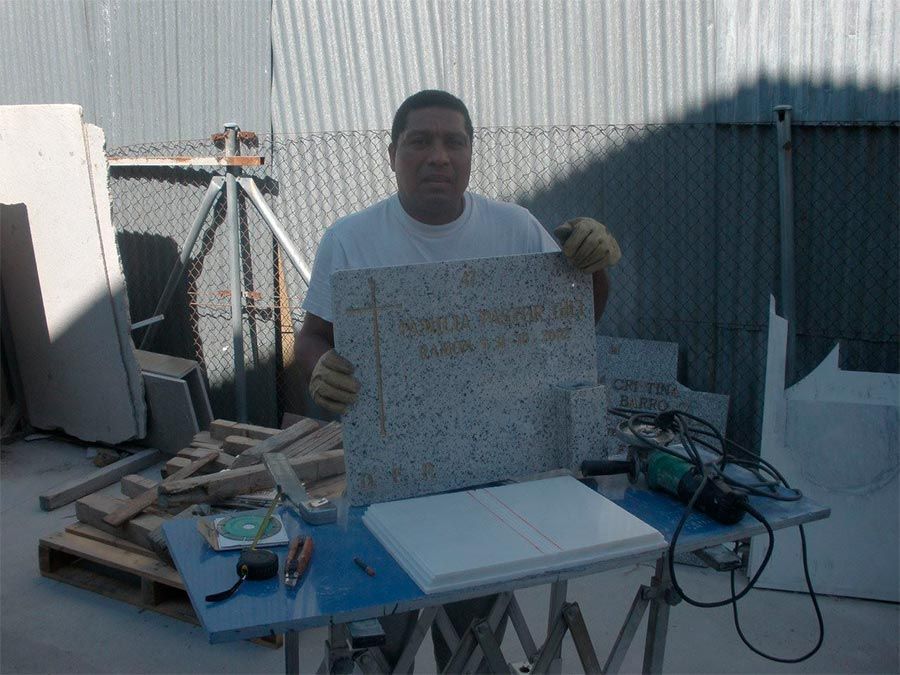 Hombre sosteniendo una pieza de granito, de pie en un banco de trabajo con herramientas, al aire libre cerca del edificio.