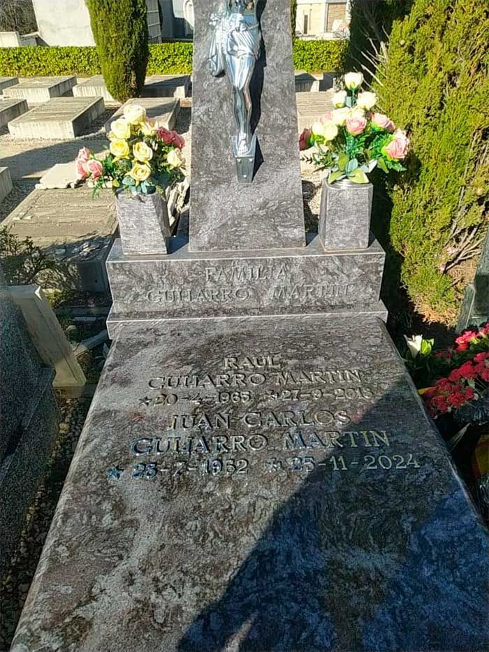 Lápida con texto y flores, una estatua de crucifijo de plata y un fondo de cementerio.