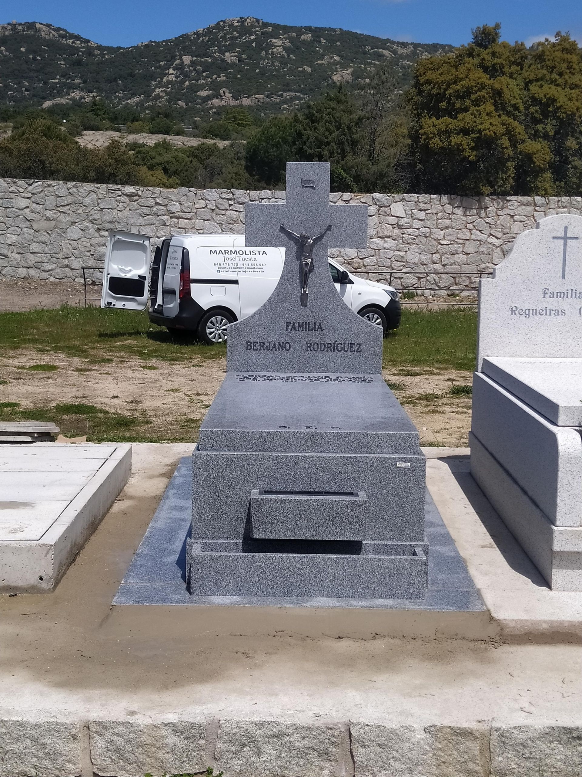 Lápida de granito con cruz y detalles esculpidos en un cementerio, con una furgoneta al fondo.