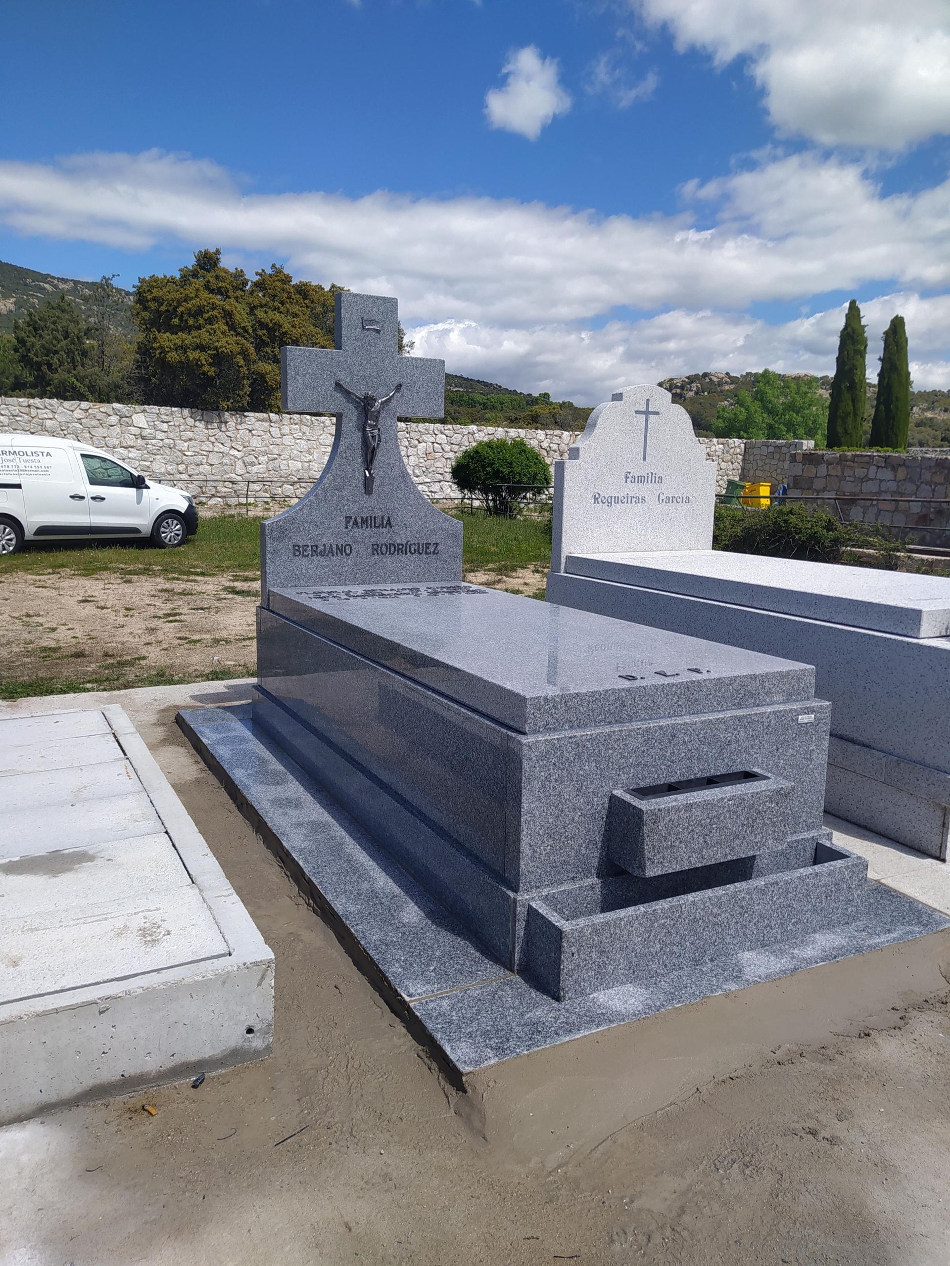 Lápida de granito con cruz, cielo azul y otras tumbas en un cementerio.