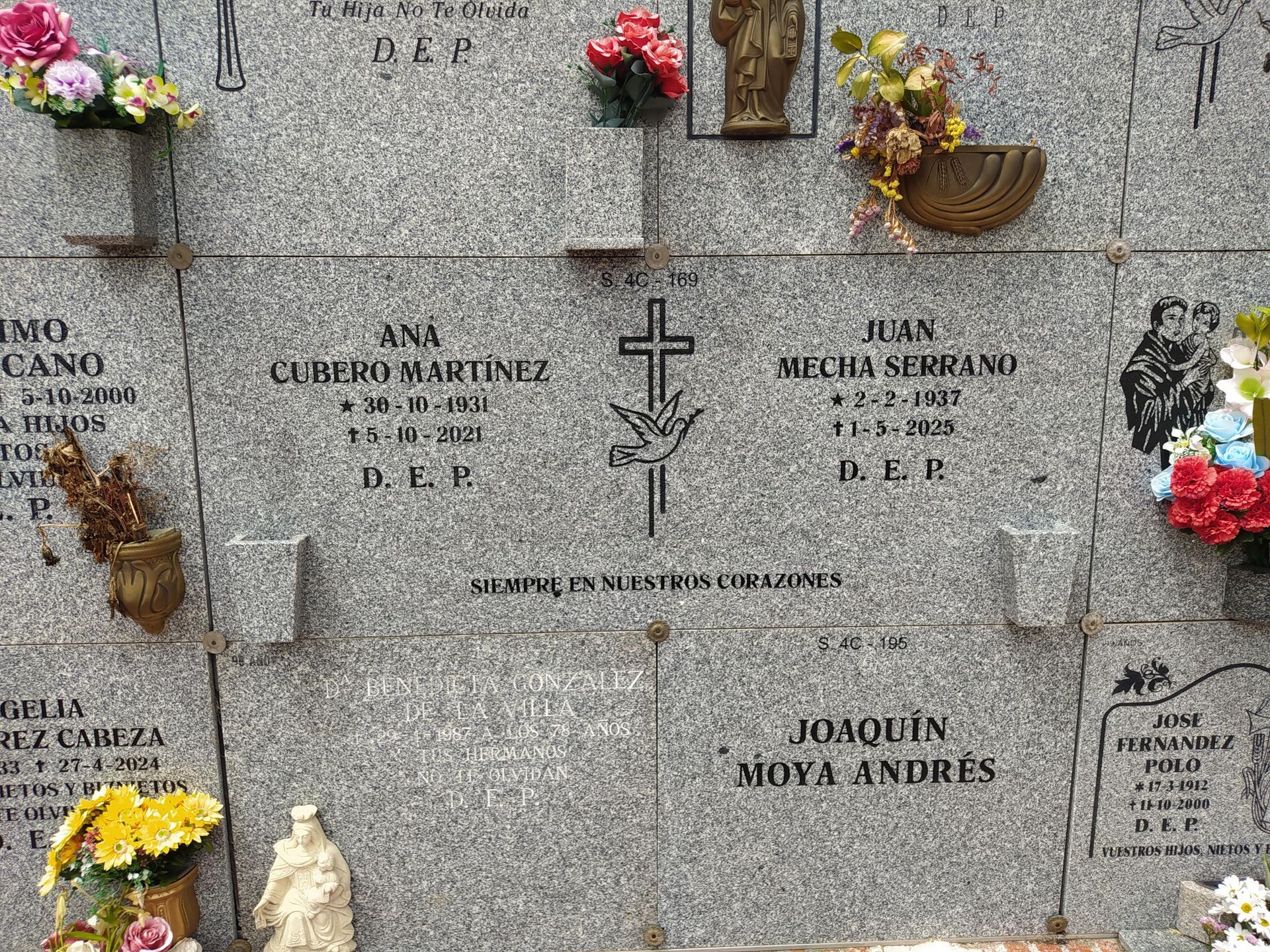 Muro de lápidas con nombres y fechas, decorado con flores y símbolo de cruz.
