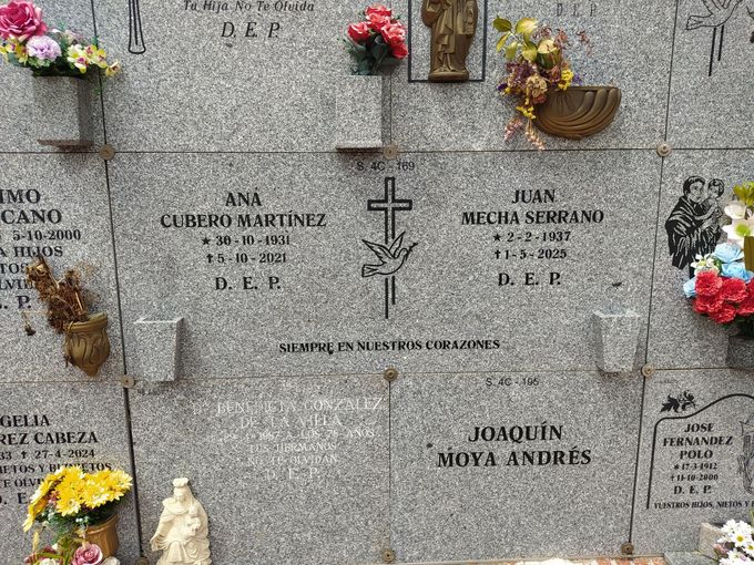 Muro de lápidas con nombres y fechas, decorado con flores y símbolo de cruz.