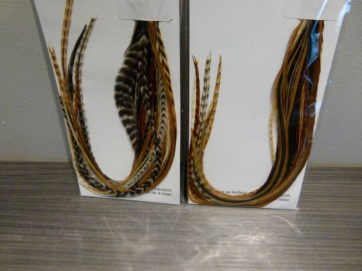 Deux extensions de cheveux à plumes dans un emballage blanc, dont une avec des plumes rayées noires et brunes.