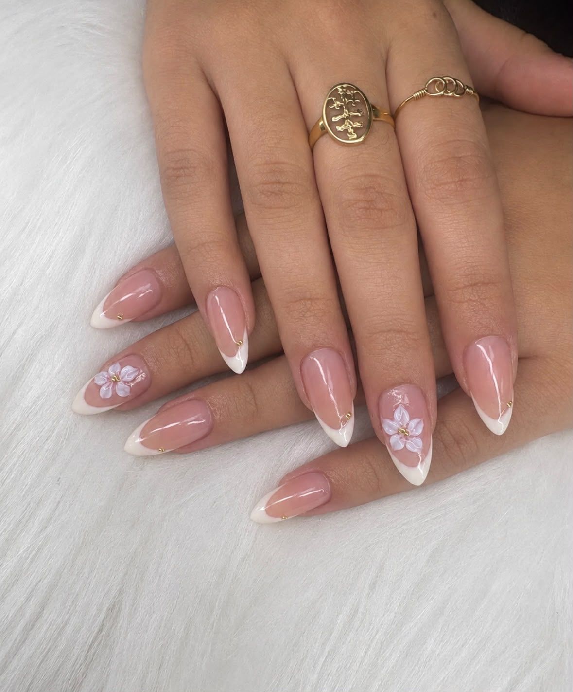 Manos con largas uñas acrílicas rosas con motivos florales y anillos dorados sobre un fondo de piel blanca.