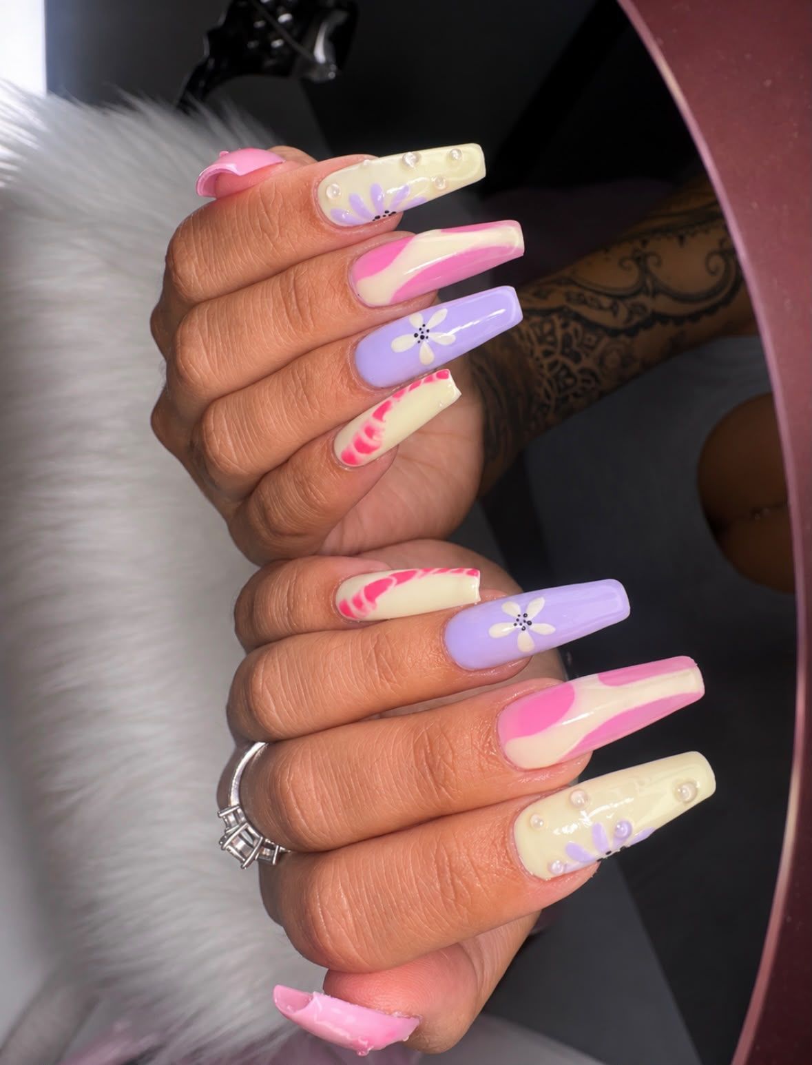 Mano con uñas acrílicas largas de color rosa pastel y lavanda, decoradas con detalles blancos y amarillos, posando sobre piel blanca.