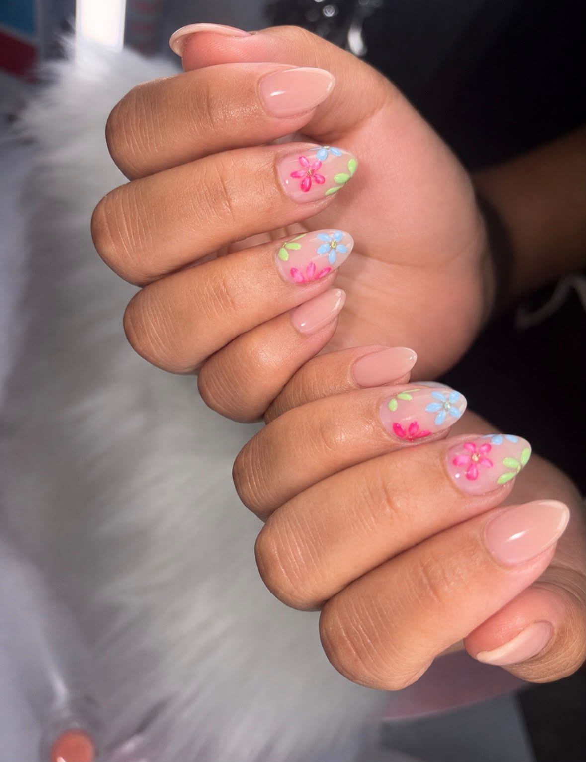 Manos con uñas largas de color rosa pastel y flores brillantes, juntas sobre un fondo blanco suave.