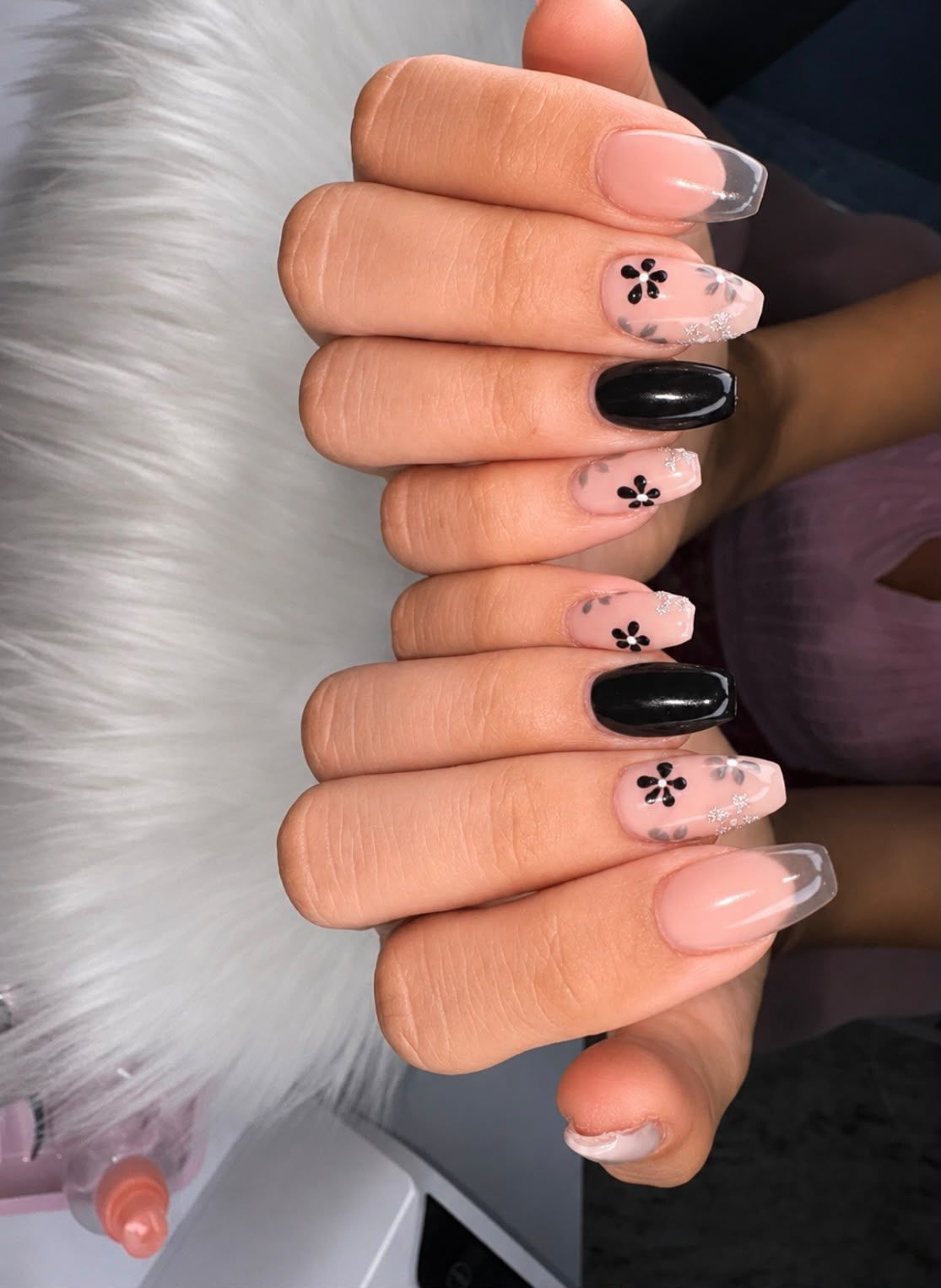 Mano con manicura floral en tonos nude y negro brillante, apoyada sobre una superficie de piel blanca.
