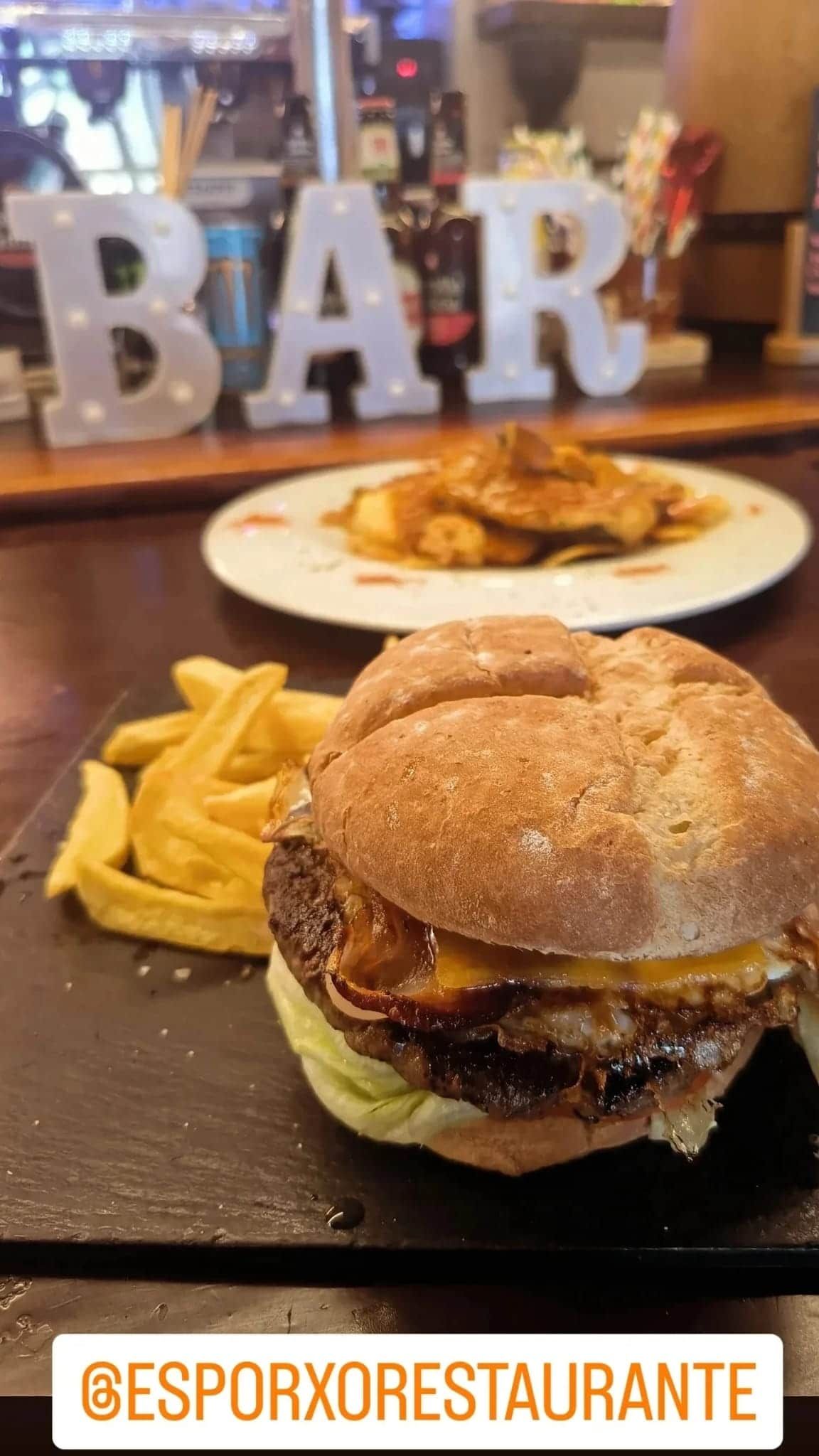 Hamburguesa con patatas fritas en un plato negro en un bar, con otro plato al fondo.