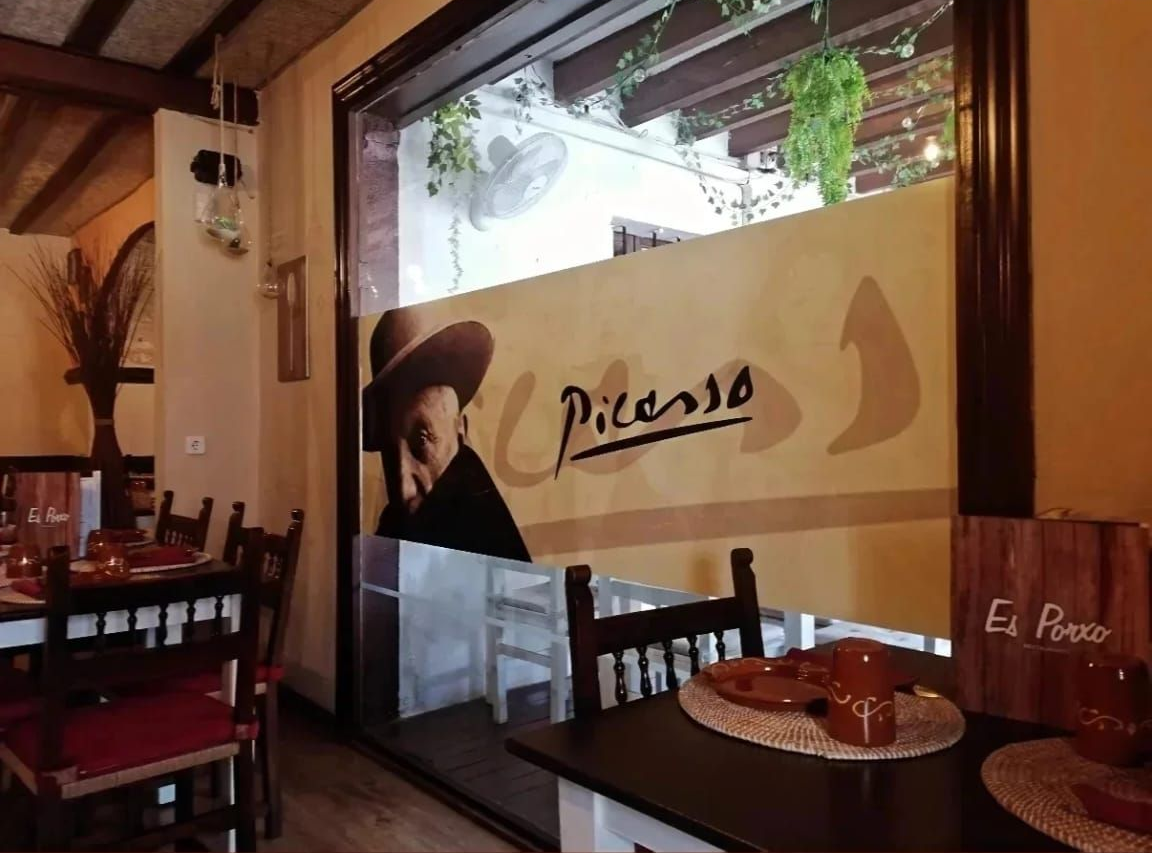 Interior de restaurante con el logo de Picasso en la ventana. Mesas y sillas de madera oscura.