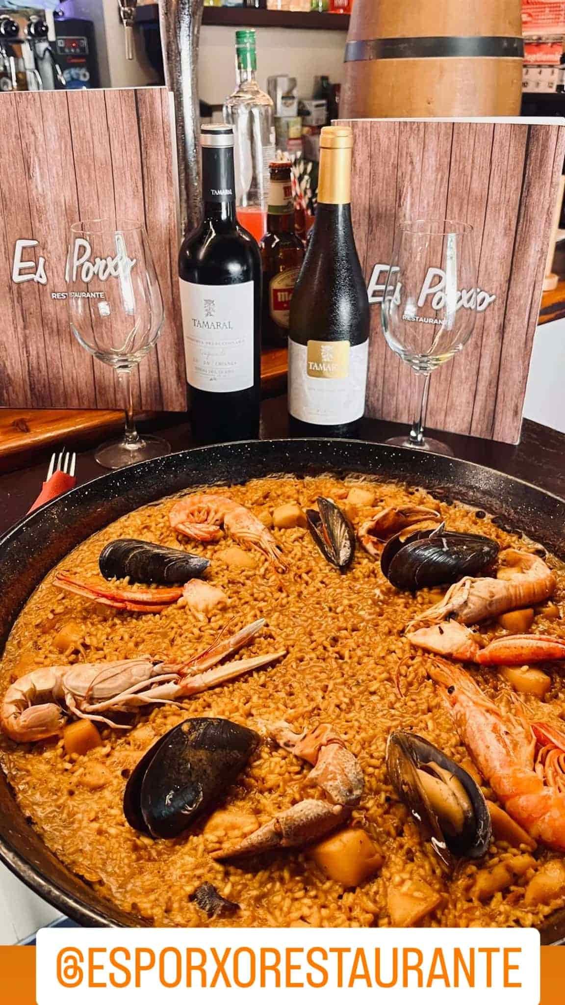 Plato de paella con marisco, botellas de vino, copas y carta en entorno de restaurante.
