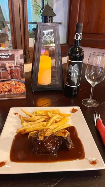 Bistec y papas fritas con salsa en un plato, una botella de vino y un vaso sobre una mesa y una linterna con vela.