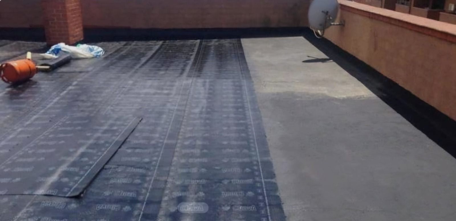 Azotea plana con membrana impermeabilizante negra y hormigón gris, junto a un muro de parapeto de ladrillo.