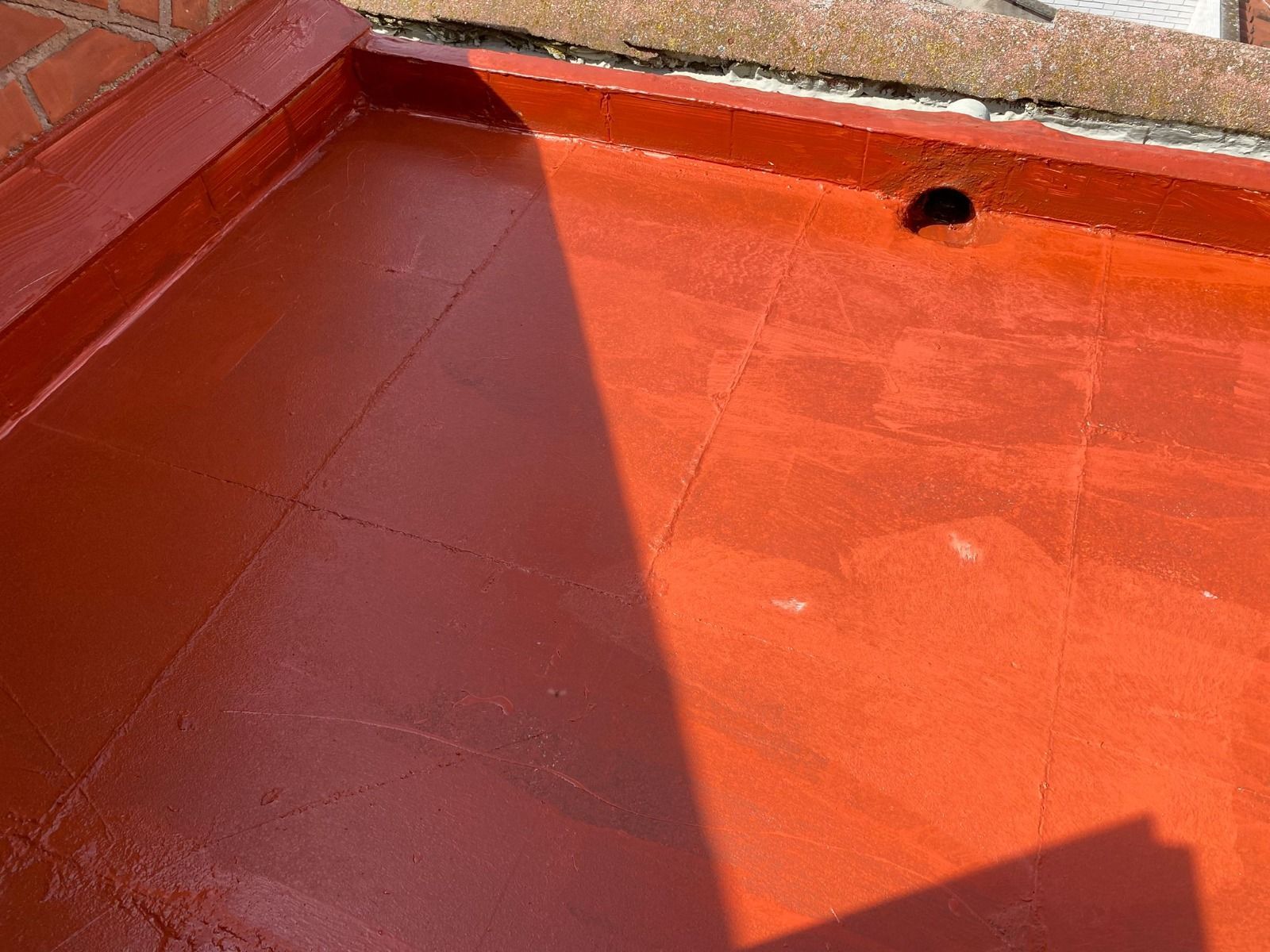 Esquina de azotea o terraza de hormigón pintada de rojo con un desagüe oscuro y una sombra en el suelo.